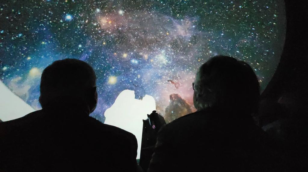 Zwei Besucher sind von hinten zusehen, welche sich die Show im Planetarium anschauen. Sie blicken auf die runde Decke des Planetariums, auf welcher ein Sternenhimmel zu sehen ist.