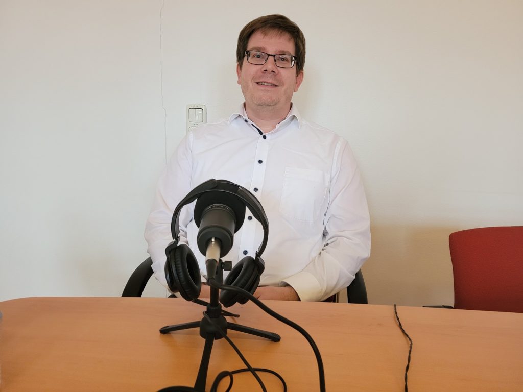 Der Geschäftsführer der Verkehrsbetriebe Hoyerswerda sitzt vor einem Mikrofon auf dem ein Paar Kopfhörer hängt und schaut in die Kamera.