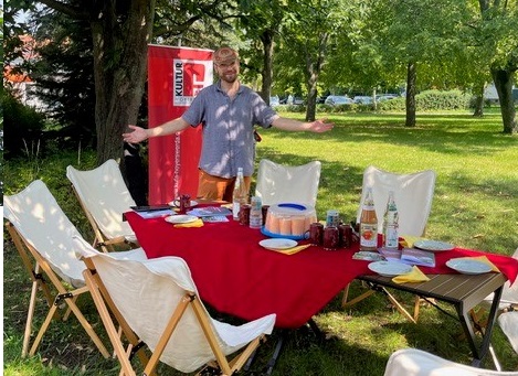 Christian Völker-Kieschnick steht mit ausgebreiteten Armen hinter einem Tisch und Stühlen, die in einem Park aufgebaut sind und auf Besucher warten.