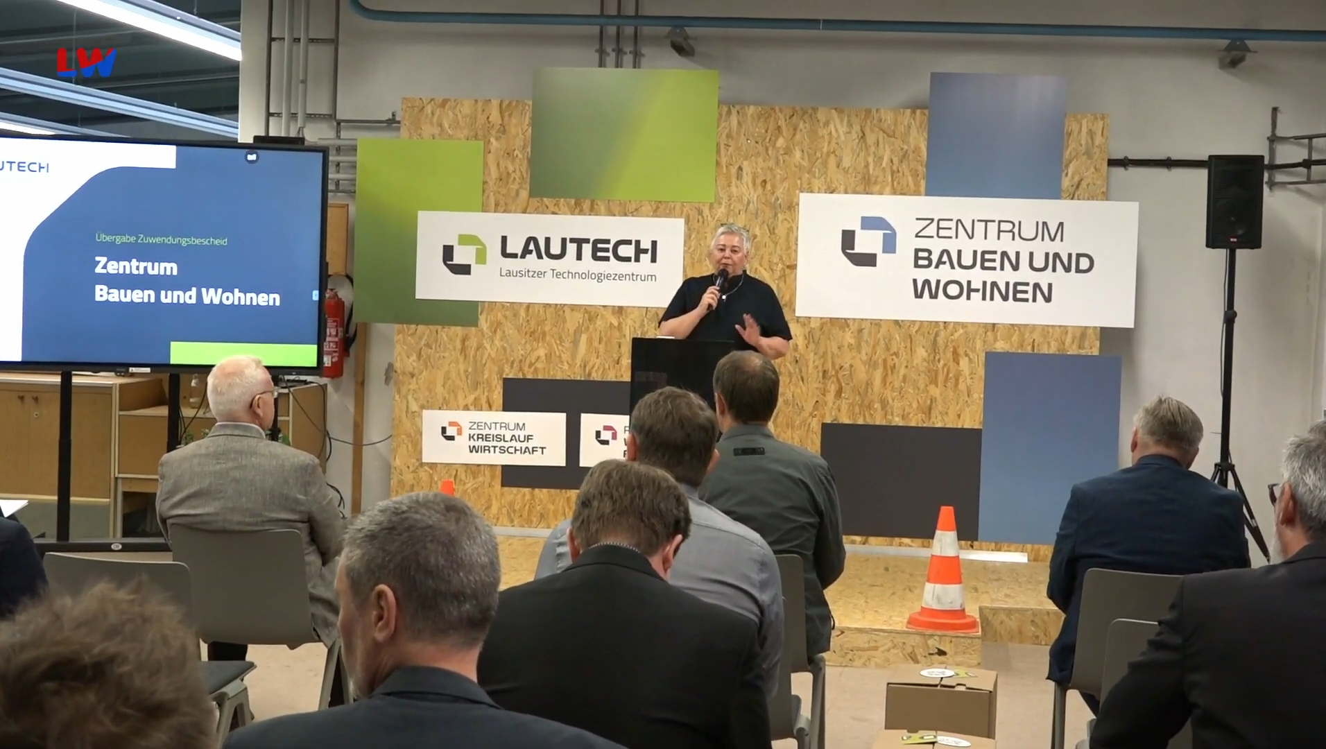 Die Geschäftsführerin der Lausitzer Technologiezentrum GmbH Kathrin Schlesinger steht auf einer Holzbühne und spricht in ein Mikrophon. Links neben ihr steht ein großer Monitor auf dem Zentrum Bauen und Wohnen steht.