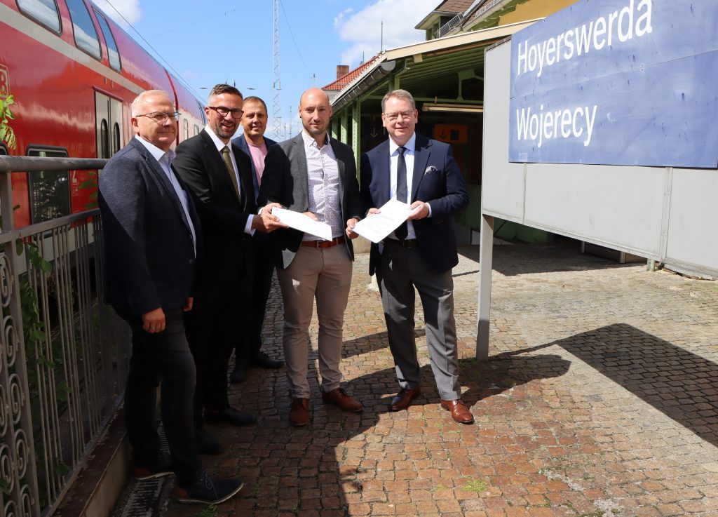 Torsten Ruban-Zeh (Oberbürgermeister der Stadt Hoyerswerda), Martin Dulig (Sächsischer Staatsminister für Wirtschaft, Arbeit und Verkehr), Mirko Pink (Bürgermeister der Stadt Hoyerswerda), Jan Riedel (DB InfraGO) und Burkhard Ehlen (Geschäftsführer ZVOE und VVO GmbH) stehen auf einem Bahnsteigt. Sie halten Fördermittelbescheid in der Hand. Links auf dem Bahngleis steht ein roter Regional Express. Rechts neben ihnen ist ein blaues Schild mit der Aufschrift Hoyerswerda sowie die sorbische Bezeichnung Wojerecy zu sehen