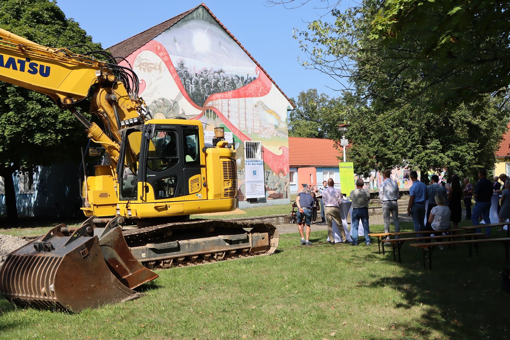 Ein großer gelber Bagger steht vor einem Gebäude. Daneben stehen rund 60 Menschen und hören zwei Menschen zu, die an einem Rednerpult stehen.