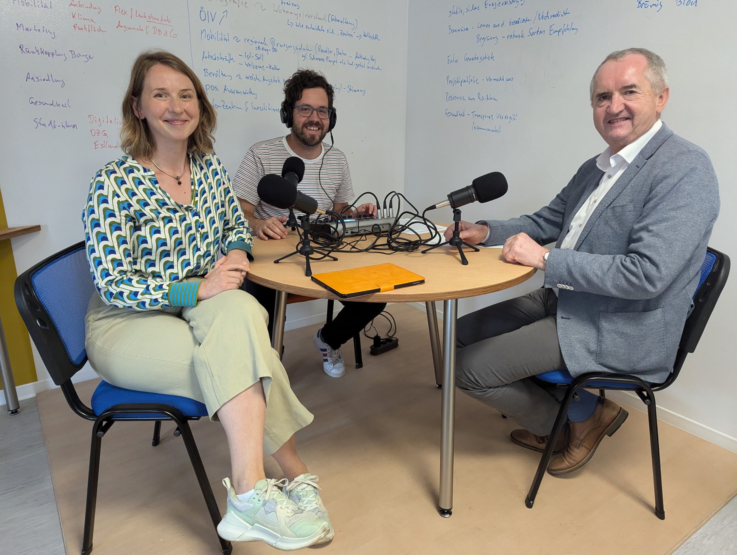 Staatsminister Thomas Schmidt, Sarah Stötzner und Robert Böhme von der Stadtverwaltung Hoyerswerda sitzen an einem kleinen runden Tisch in einem Raum und lächeln in die Kamera. Auf dem Tisch stehen ein Mischpult und drei Mirkofone.