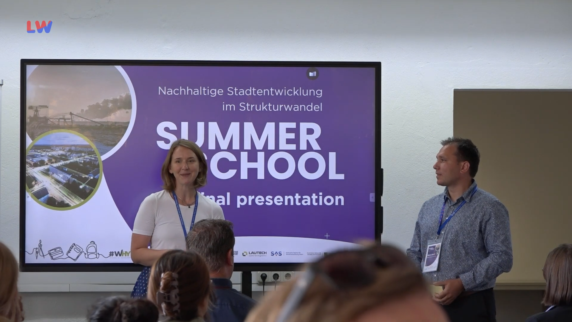 Sarah Stötzner und Marco Bloch vom Strukturwandelteam der Stadt Hoyerswerda stehen vor einer digitalen Tafel und begrüßen die etwa 30 Gäste bei der Präsentationsveranstaltung der Summer School für nachhaltige Stadtentwicklung.