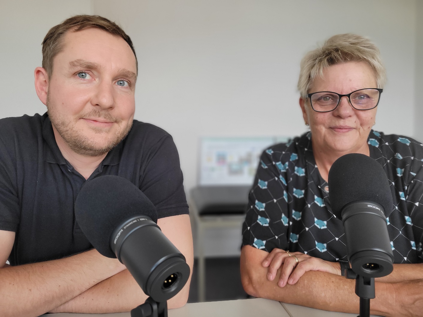 Jens Leschner und Evelyn Scholz von der RAA Ostsachsen Hoyerswerda e.V. sitzen nebeneinander an einem Tisch und lachen in die Kamera. Vor ihnen stehen zwei Mikrofone.